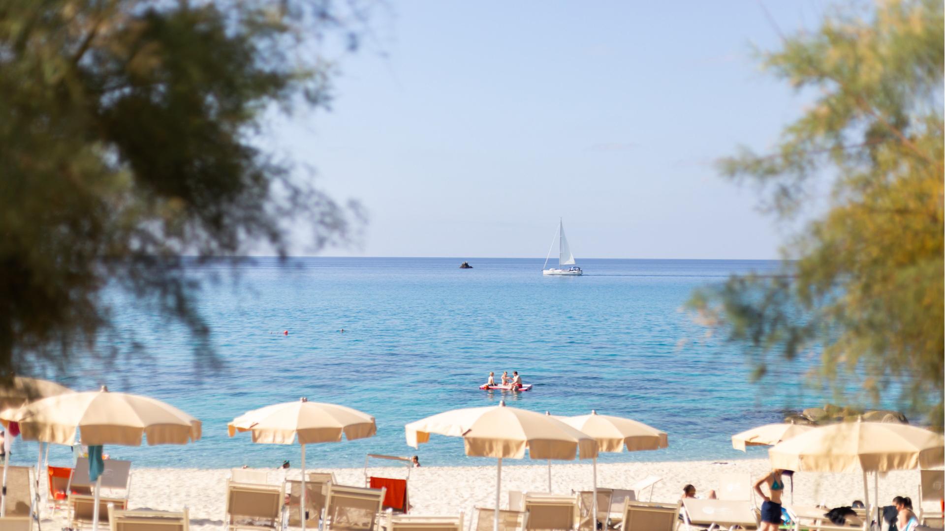 Plage de sable avec parasols, mer bleue et voilier à l'horizon.