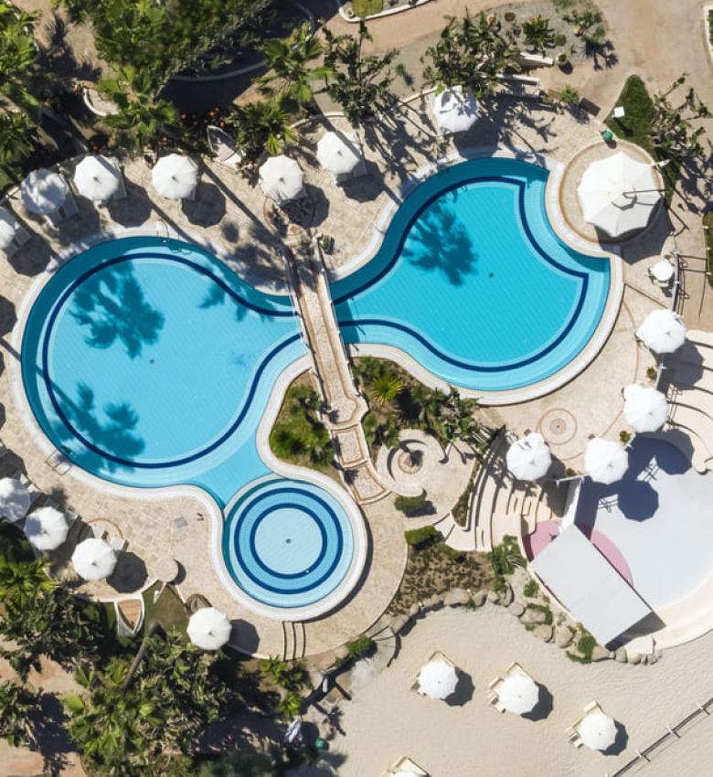 Luftaufnahme eines Pools mit Sonnenschirmen und Palmen.